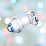 A rounded glass piece with a bulbous tip and flat base showcases swirling blue patterns, reminiscent of the Gläs Pleasure Droplets Anal Training Kit. The background highlights a blurred bokeh effect of soft multicolored lights.