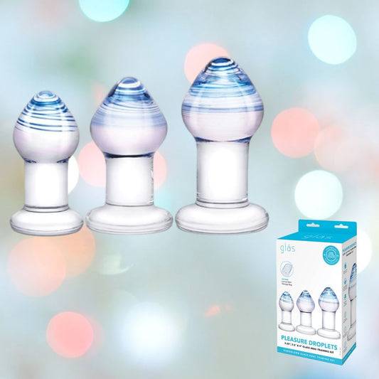 Against a colorful backdrop, three elegant glass droplets feature rounded tops with a blue swirl design. A box labeled "Gläs Pleasure Droplets Anal Training Kit" suggests they are perfect for both anal training and use as versatile glass butt plugs. 1080