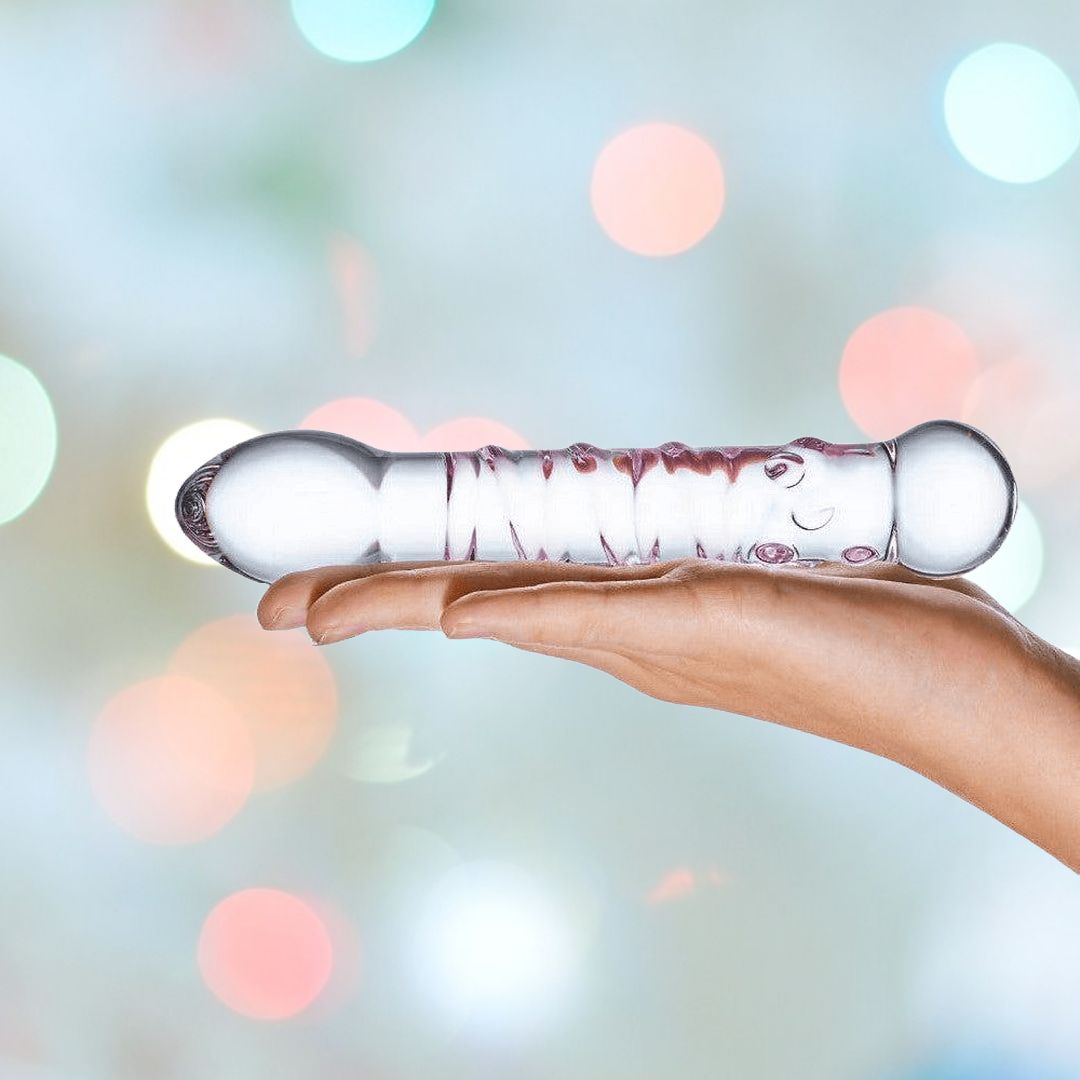 A hand holds the Gläs Spiral Staircase Full Tip G-Spot Glass Dildo against a blurred background with pastel-colored bokeh lights. The clear dildo, adorned with pink swirls inside, features a bulbous end and a rounded end, making it perfect for G-spot massage.