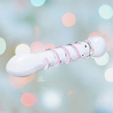 A Gläs Spiral Staircase Full Tip G-Spot Glass Dildo with a smooth, rounded, bulbous end, wrapped in light pink bands along its length. The blurred, colorful bokeh background adds a festive or dreamy element to the scene.