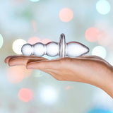 A hand holding a Gläs Titus Glass Beaded Butt Plug Dildo with a bulbous design against a blurred background featuring soft pastel-colored bokeh lights.