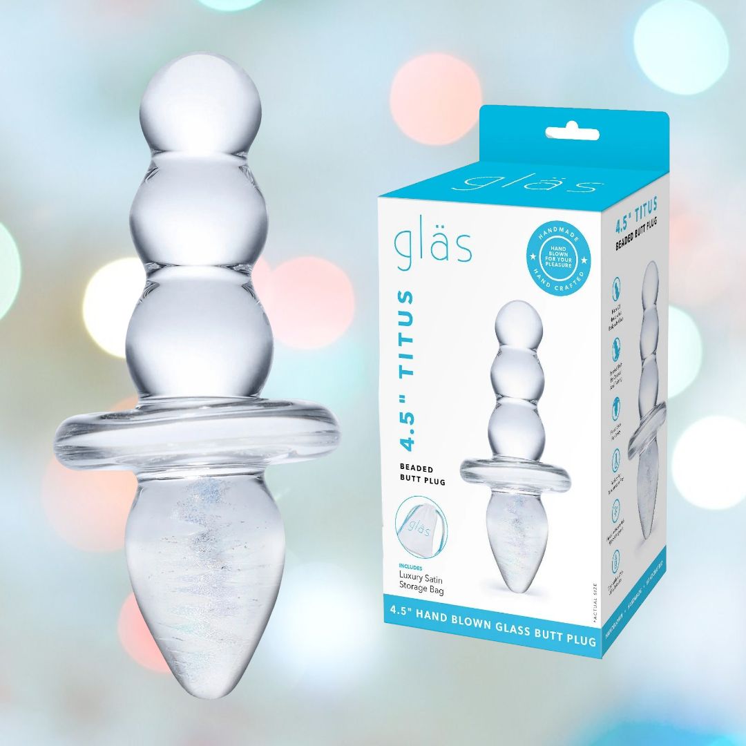 The Gläs Titus Glass Beaded Butt Plug Dildo, featuring three rounded sections for gentle anal dilation and a flared base, is displayed next to its packaging labeled "Gläs 4.5'' Titus." The background features soft-focus pastel colors that emphasize the hypoallergenic properties of this sophisticated product.