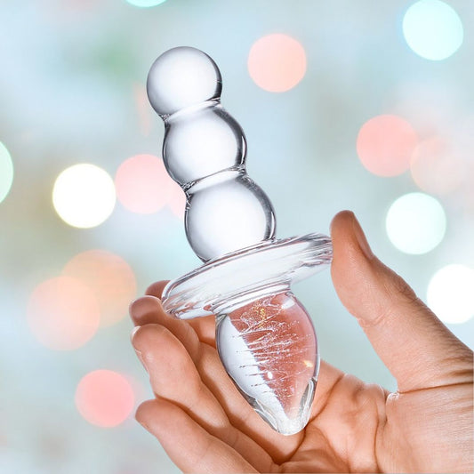 A hand holding a Gläs Titus Glass Beaded Butt Plug Dildo, featuring multiple spherical sections, against a softly blurred background with pastel-colored bokeh lights. 1080