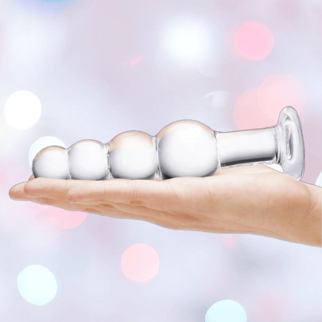 A hand holds the Gläs 7.25" Luxury Glass Beaded Butt Plug by Gläs, featuring four round graduated beads and a flared base—crafted from body-safe glass for temperature play, shown against a soft pastel bokeh background.