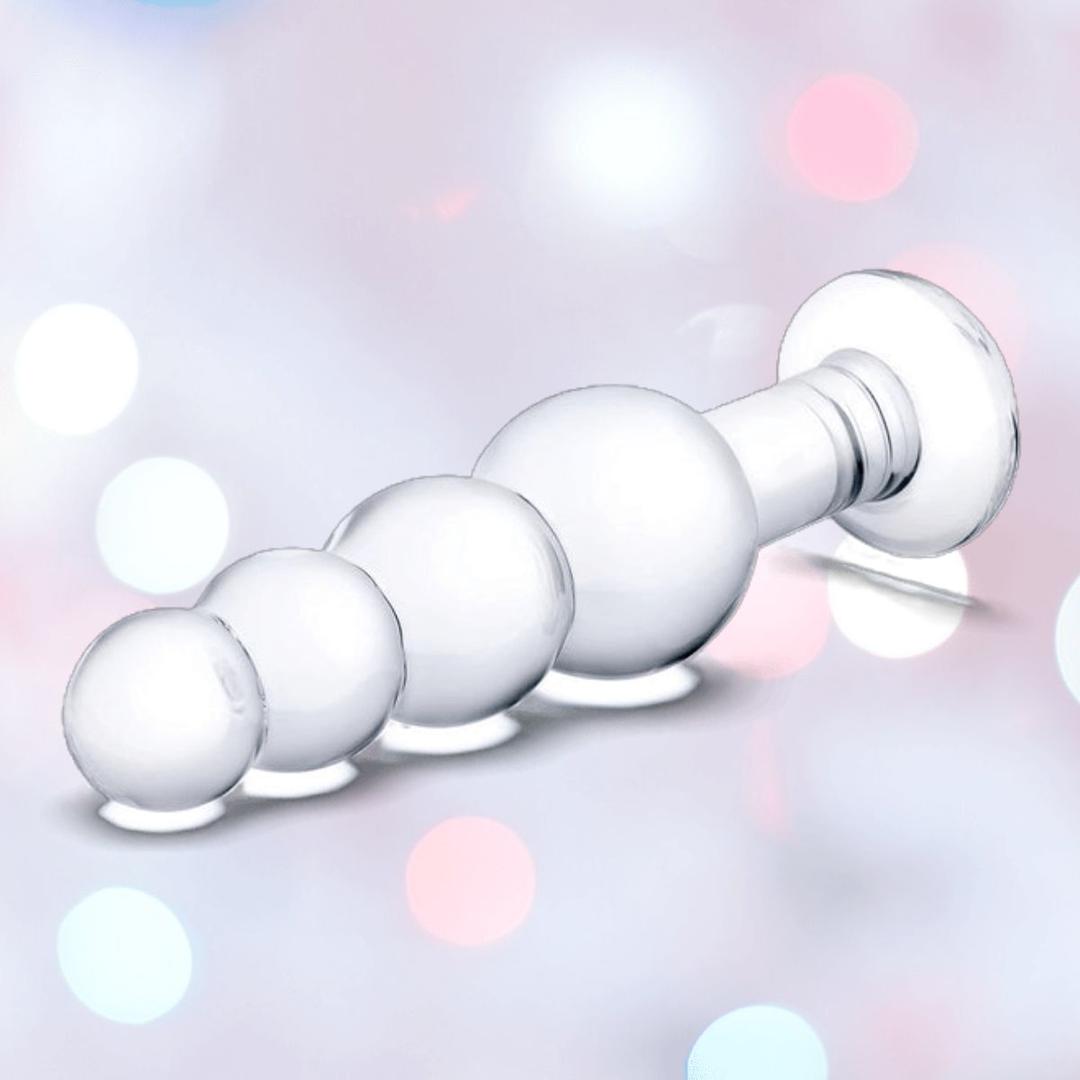 The Gläs 7.25" Luxury Glass Beaded Butt Plug by Gläs is shown on a softly blurred pastel background with bokeh lights, highlighting its body-safe glass ideal for temperature play and four graduated spheres with a flared base.
