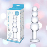 The Gläs 7.25" Luxury Glass Beaded Butt Plug, made from body-safe glass with a flared base, is shown next to its white and blue Gläs packaging. Soft pastel pink and blue tones in the background enhance its inviting look for temperature play.