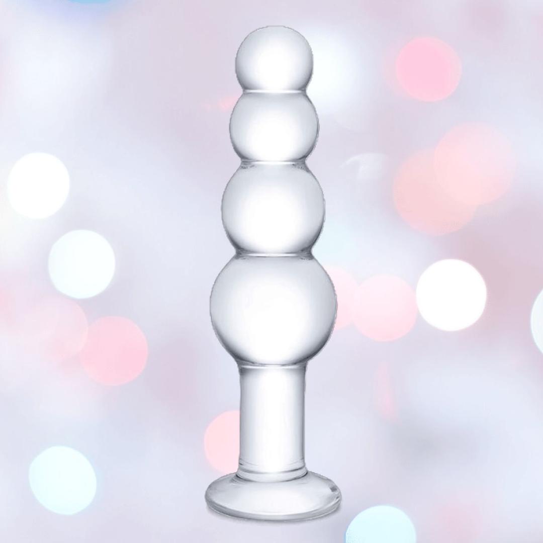 The Gläs 7.25" Luxury Glass Beaded Butt Plug by Gläs features four smooth, stacked bulbs in body-safe clear glass, displayed against a pastel bokeh background with pink and white lights.