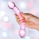 A hand holds the Gläs Double Trouble Luxury Dual-Head Glass Dildo - Purple by Gläs against a softly blurred pastel background with circular light spots.
