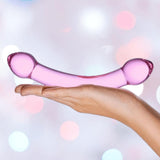 A hand holds the Gläs Double Trouble Luxury Dual-Head Glass Dildo in purple, its curved, translucent form and rounded ends set against a softly blurred pastel light background.