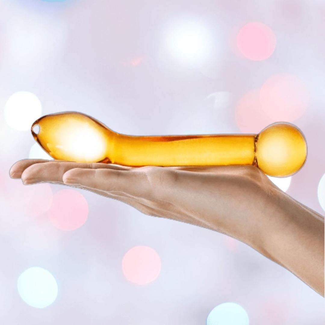 A hand holds the Gläs Honey Dripper 7" Double Ended Glass Anal and G-Spot Dildo, featuring a transparent amber hue with rounded and bulbous tips, set against a softly blurred pastel background.
