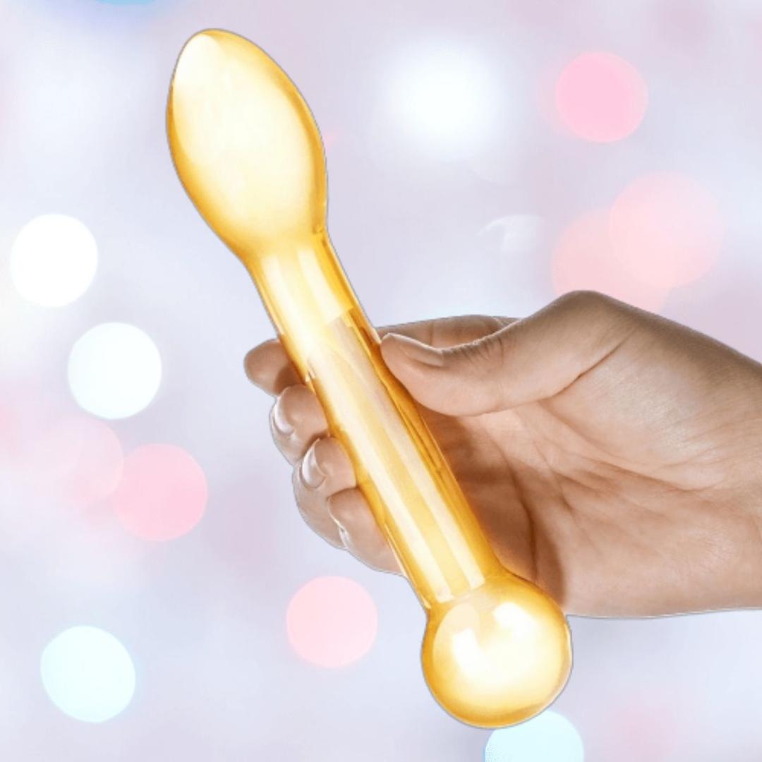 A hand holds the Gläs Honey Dripper 7" Double Ended Glass Anal and G-Spot Dildo by Gläs, a translucent yellow toy with a rounded ball end and flat spoon-like tip, ideal for P-Spot or G-Spot play, against soft colorful lighting.