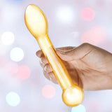 A hand holds the Gläs Honey Dripper 7" Double Ended Glass Anal and G-Spot Dildo by Gläs, a translucent yellow toy with a rounded ball end and flat spoon-like tip, ideal for P-Spot or G-Spot play, against soft colorful lighting.