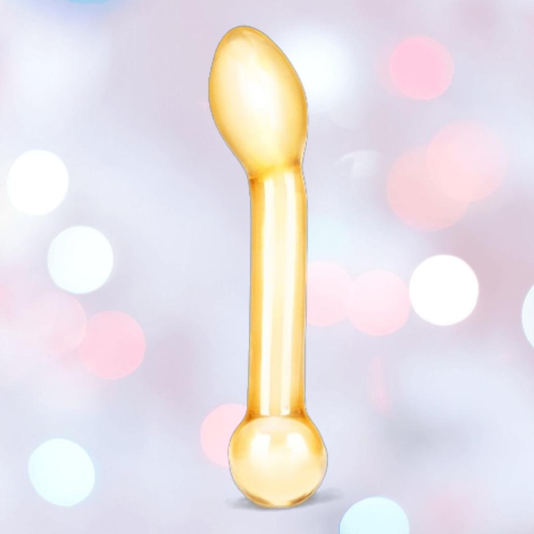 The Gläs Honey Dripper 7" Double Ended Glass Anal and G-Spot Dildo by Gläs, featuring a yellow-tinted, bulbous design with a rounded angled head, is displayed against a soft pink, white, and blue bokeh background.