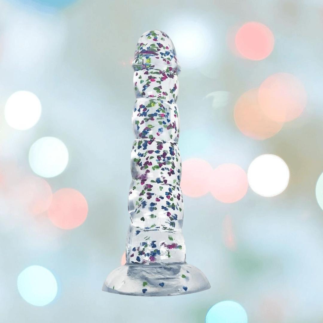The Glittergasm Dong - Blue and Green Swirl 7" by Icon Brands stands upright, highlighting its premium silicone build, colorful swirled design, and harness compatibility against a softly blurred pastel background.