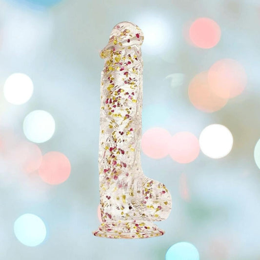 The Icon Brands Glittergasm Dong - Red and Orange Swirl 7" is a transparent, phallic-shaped dildo with colorful confetti inside, a suction base, and harness compatibility, shown against a pastel backdrop with soft circular lighting. 1080