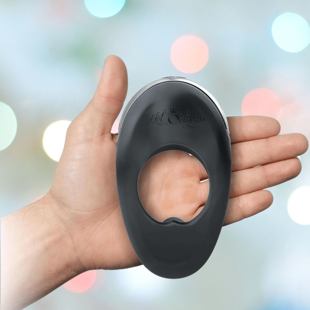 Close-up of a hand holding the Hot Octopuss Atom Plus - Vibrating Cock Ring in black silicone with "Hot Octopuss" embossed on it. The background is blurred with colorful bokeh lights, hinting at its powerful vibration designed for perineum stimulation.