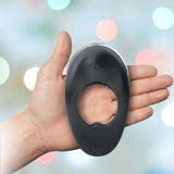 Close-up of a hand holding the Hot Octopuss Atom Plus - Vibrating Cock Ring in black silicone with "Hot Octopuss" embossed on it. The background is blurred with colorful bokeh lights, hinting at its powerful vibration designed for perineum stimulation.