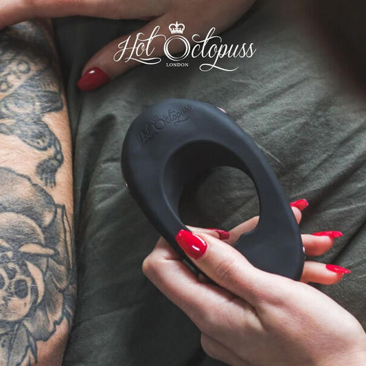 A person holds a black Hot Octopuss Atom Plus, a vibrating cock ring known for its powerful vibrations, while another with a tattooed arm lies nearby on a bed with green sheets. The words "Hot Octopuss London" are visible at the top of the image. 1080