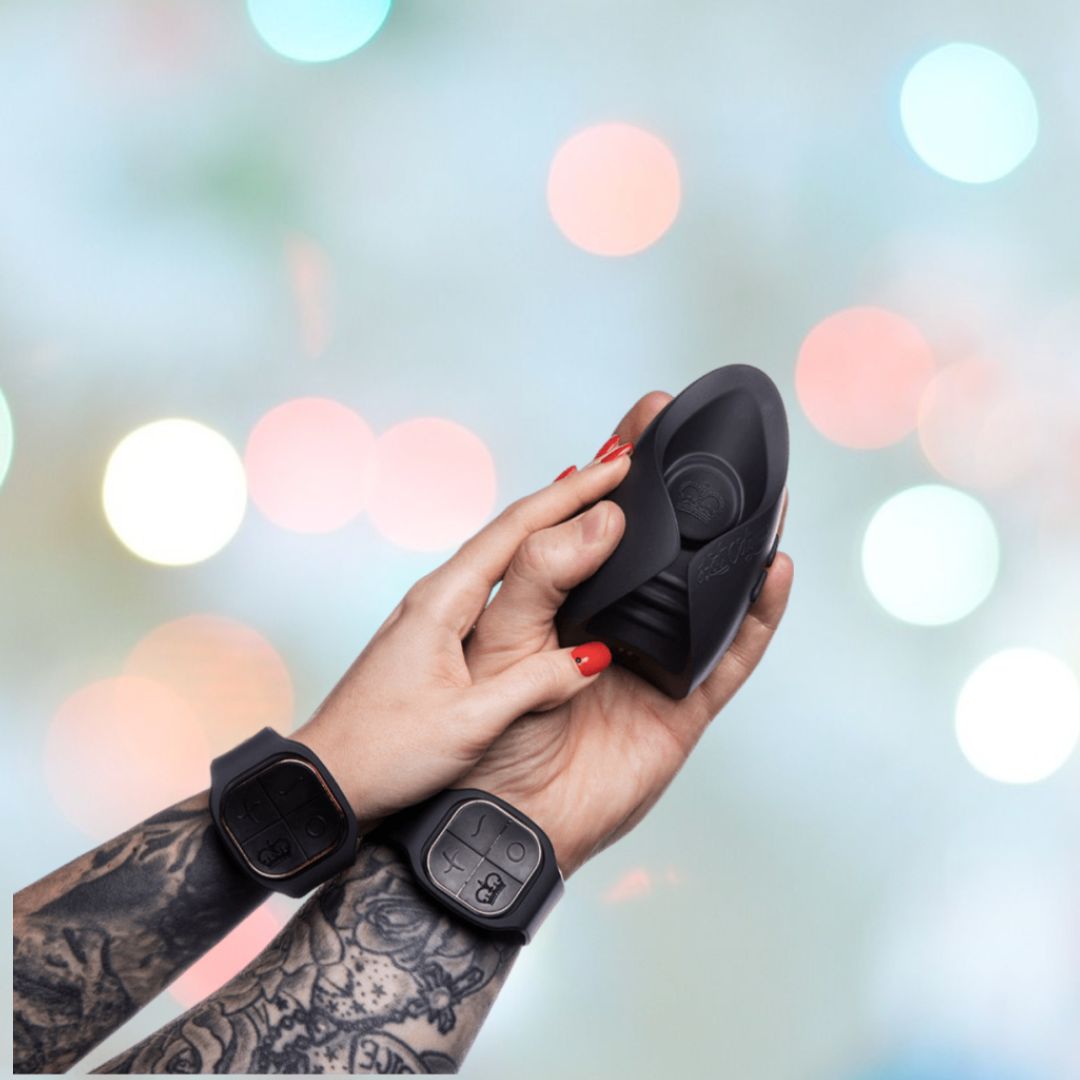 A person with tattoos and red nail polish is holding the sleek black Hot Octopuss Pulse DUO LUX Couples Vibrator against a blurred background featuring colorful lights. Both wrists are adorned with black watches with large dials, enhancing the futuristic allure.