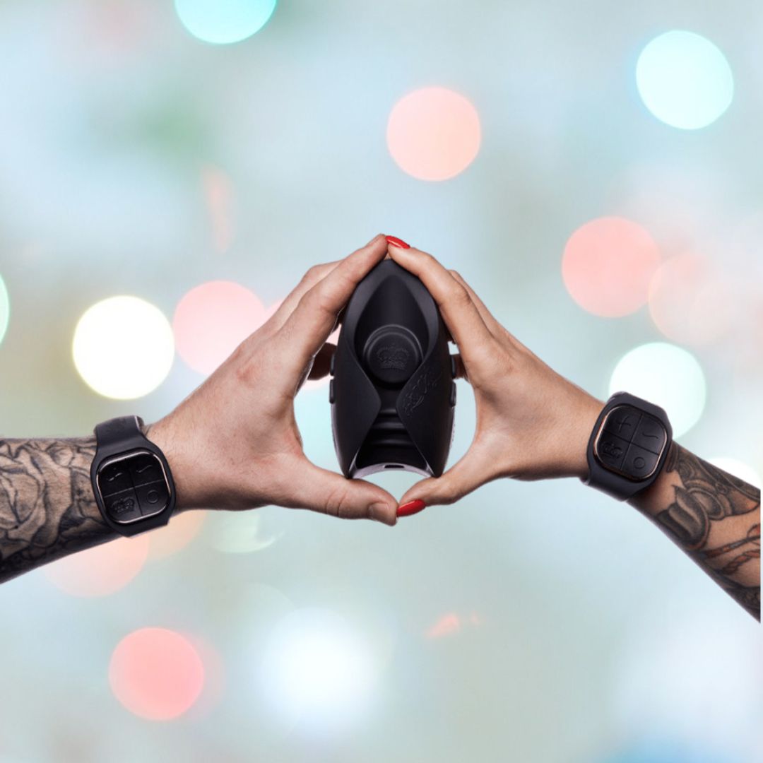 Two tattooed hands wearing black smartwatches form a heart shape around a sleek black bottle of the Hot Octopuss Pulse DUO LUX Couples Vibrator. The background, adorned with colorful bokeh lights, enhances the enchanting vibe of this special moment with the couple's toy from Hot Octopuss.