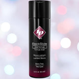 A 2.2 fl oz (65 ml) black bottle of ID Lubricants' ID Backslide Silicone Anal Lubricant, labeled extra thick, stands against a backdrop of blurred pink and white lights.