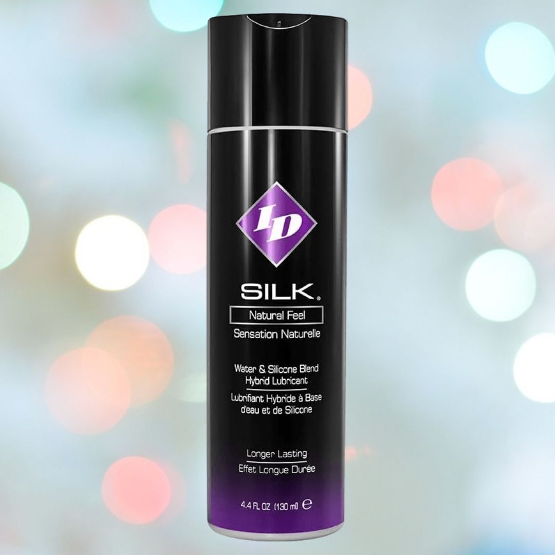 A black and purple bottle of ID Silk Long-Lasting Hybrid Lubricant from ID Lubricants is set against a blurred background with colorful light bokeh. The bottle, labeled as a long-lasting water and silicone blend hybrid lubricant, holds 4.4 fl oz (132 ml).