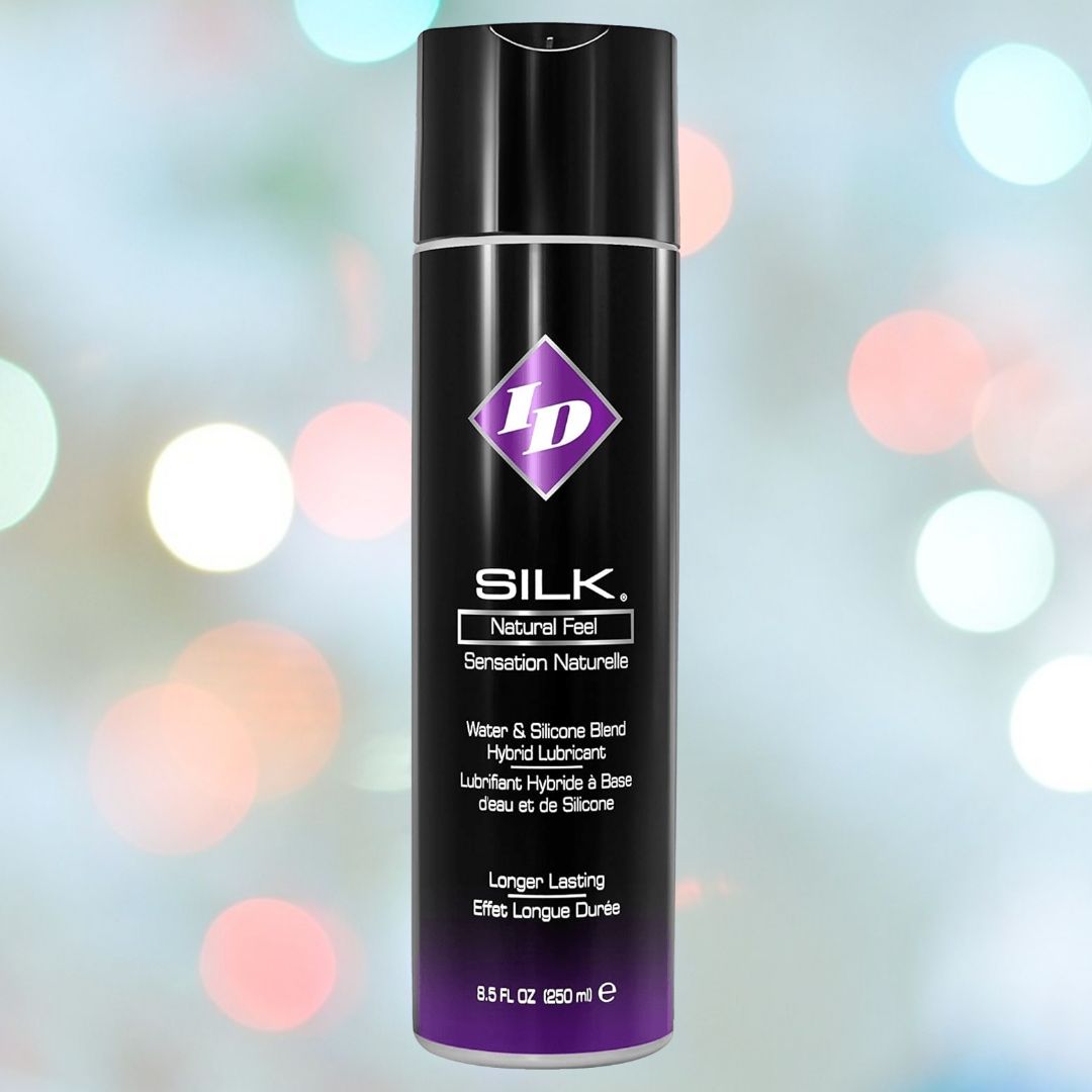 Image of a can of ID Silk Long-Lasting Hybrid Lubricant by ID Lubricants, featuring a black and metallic purple design with white text detailing it as a water and silicone blend for a natural feel. The background is softly blurred with pastel-colored bokeh lights. Contains 8.5 fl oz (250ml).