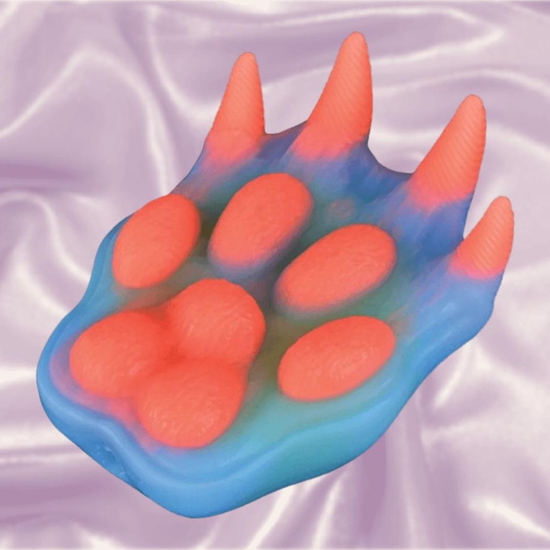 The Icon Brands Alien Nation Pantera Grinder, by Icon Brands, is a silicone grinder shaped like a colorful animal paw with four raised pads and claws in blue and pink-orange hues, glowing in the dark on light purple fabric.