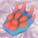 The Icon Brands Alien Nation Pantera Grinder, by Icon Brands, is a silicone grinder shaped like a colorful animal paw with four raised pads and claws in blue and pink-orange hues, glowing in the dark on light purple fabric.