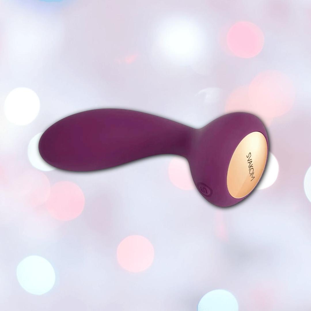 The Svakom JULIE Wearable Anal & G-Spot Vibrator with Remote Control, featuring a purple and gold elongated body with a rounded base, is shown against a softly blurred pastel bokeh background.
