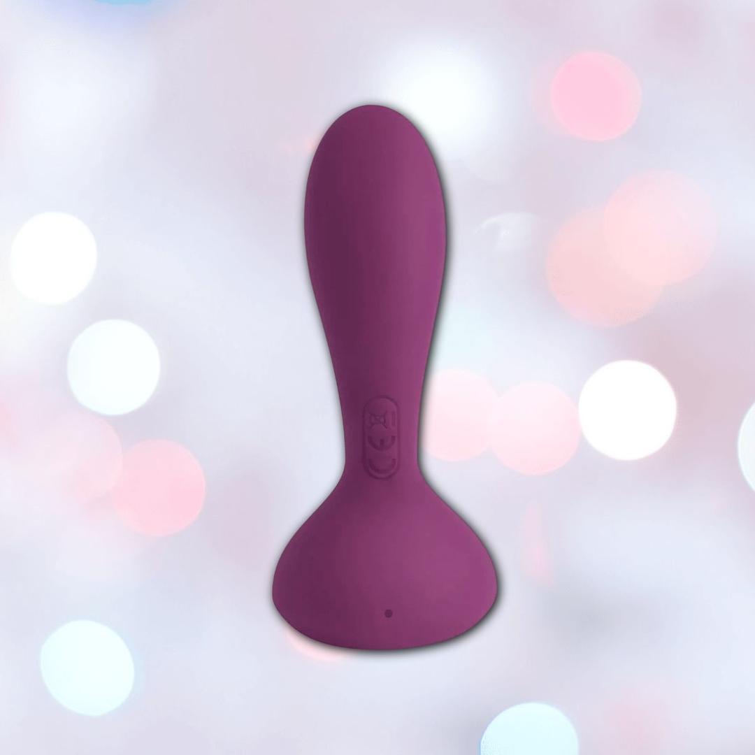 The Svakom JULIE Wearable Anal & G-Spot Vibrator with Remote Control, in purple silicone with a flared base and ergonomic shape, is displayed against a soft, blurred pastel background featuring light pink and white bokeh.