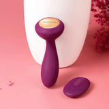 A Svakom JULIE wearable anal & G-spot vibrator with a gold accent and matching remote control rests on a pink surface beside a white vase and red flowers.