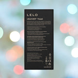 A box labeled "LELO DOT Travel Clitoral Pinpoint Vibrator Pink" is set against a blur of colorful lights. Featuring Infinite Loop Technology, this clitoral vibrator details its features in white text, with logos and contact info at the bottom.