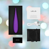 The image displays a boxed LELO DOT Travel Clitoral Pinpoint Vibrator in purple, highlighting its Infinite Loop Technology, and includes a warranty card, USB charging cable, black storage pouch, and instruction manual. It's an ideal travel companion set against a colorful bokeh background.