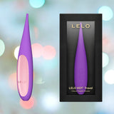 The LELO DOT Travel Clitoral Pinpoint Vibrator in purple is elegantly presented next to its sleek black box packaging. The backdrop of soft, colorful bokeh lights highlights its appeal as an ideal travel companion.