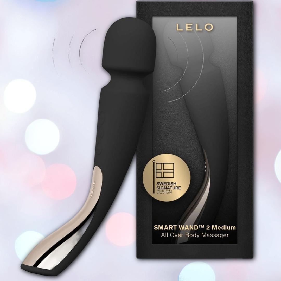 The LELO Smart Wand 2 Medium Personal Wand Massager by LELO in black is displayed beside its box, which features a gold emblem and product name. Soft, blurred pastel lights in the background highlight this stylish waterproof wand.