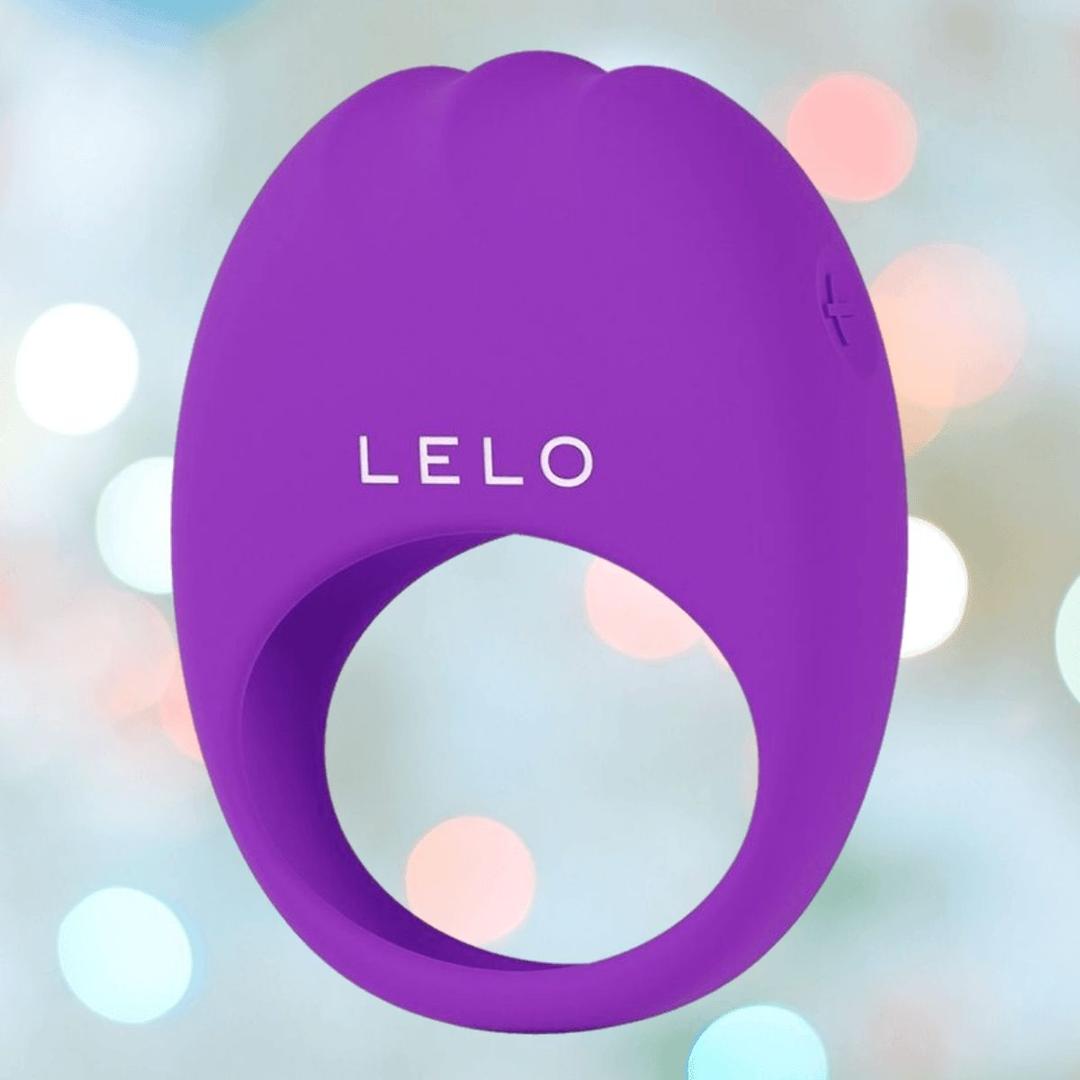 The LELO LIFEGUARD Originals Purple, a silicone ring-shaped device with "LELO" branding, appears against a softly blurred background featuring pastel tones and circular light spots.