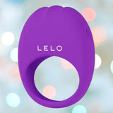 The LELO LIFEGUARD Originals Purple, a silicone ring-shaped device with "LELO" branding, appears against a softly blurred background featuring pastel tones and circular light spots.