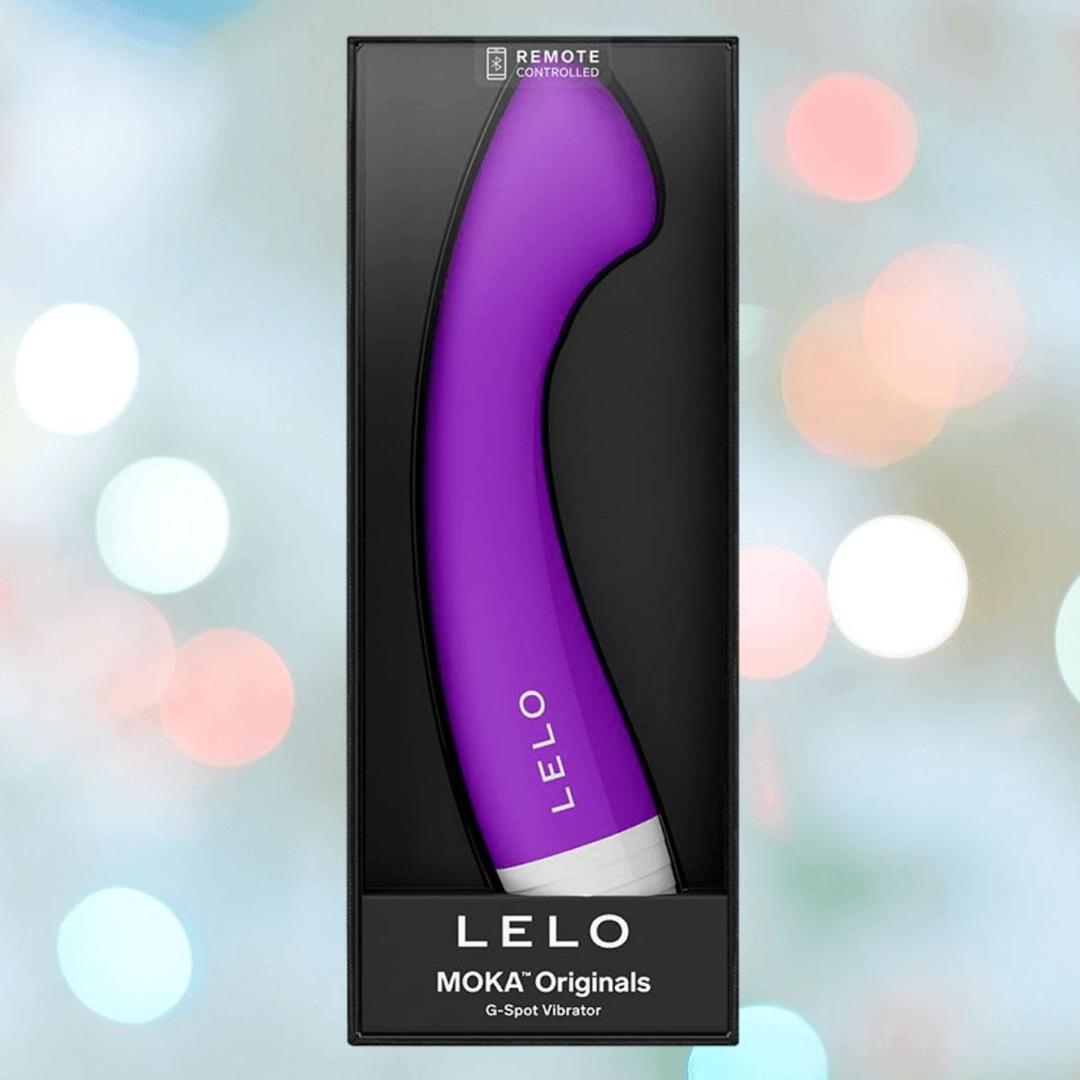A Lelo MOKA Originals Purple G-spot vibe by LELO stands upright in its black box, with the brand name at the bottom, set against a blurred pastel background. This sleek vibrator is waterproof and comes in stylish retail packaging.