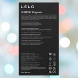 The back of a LELO SURFER Originals Purple box features multilingual descriptions and instructions against a blurred pastel bokeh background, with LELO branding and certification logos displayed at the bottom.