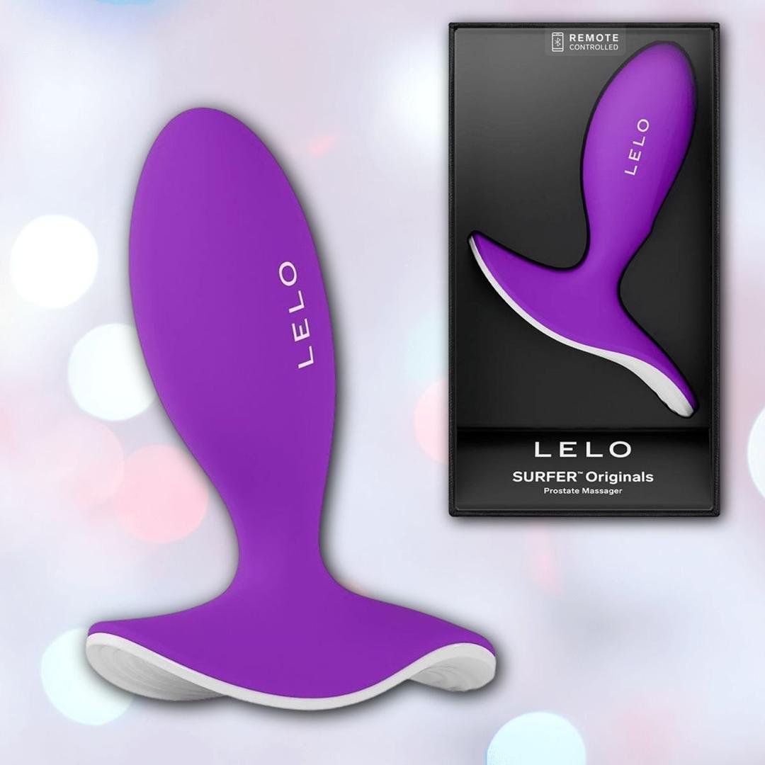 The Lelo SURFER Originals Purple by LELO is displayed beside its black packaging, which showcases the product and brand name. The scene features a softly blurred bokeh background.