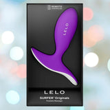 The LELO SURFER Originals Purple prostate massager is shown in sleek black packaging, featuring remote control text. The softly blurred background with light bokeh highlights the stylish LELO design.