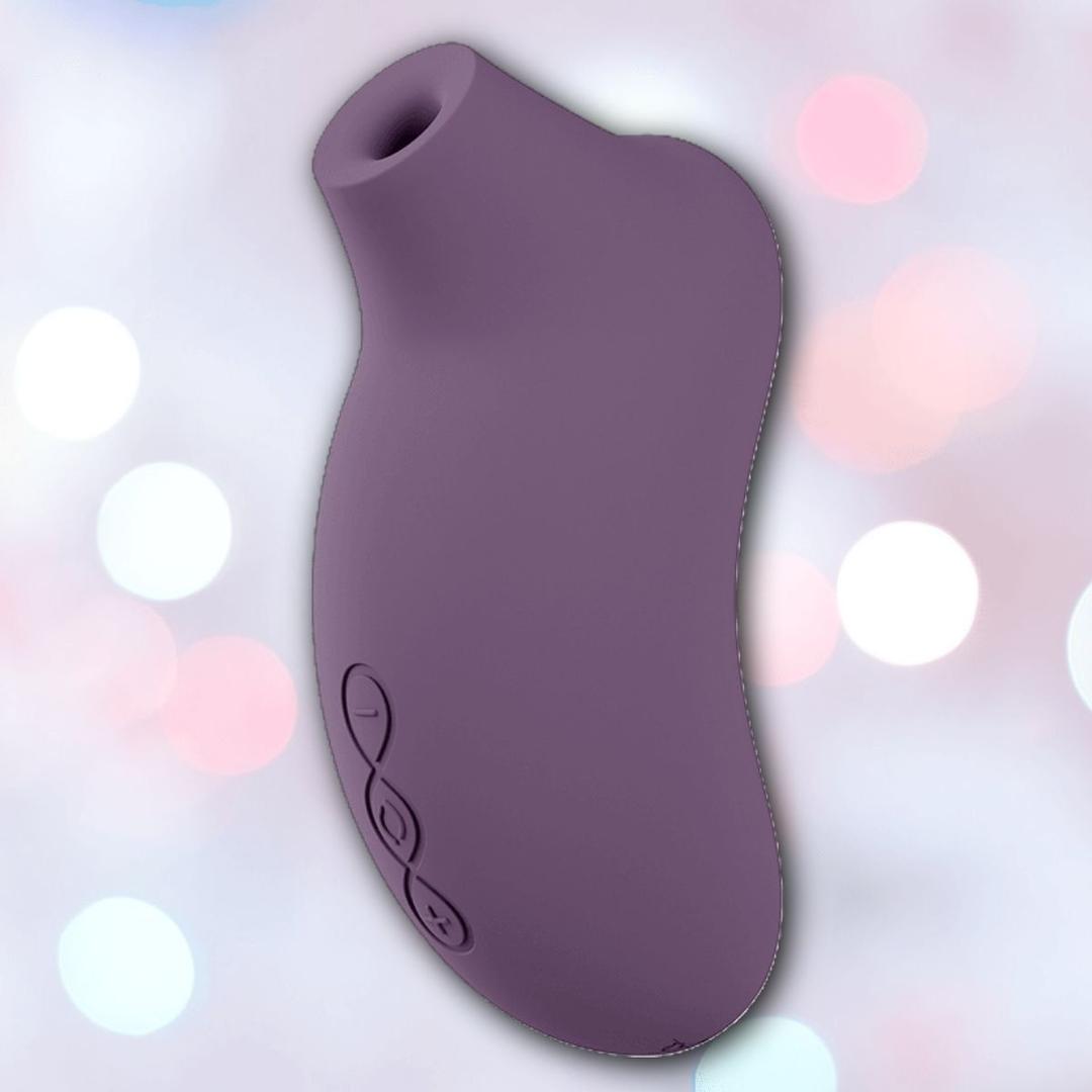 The LELO Sona 3 Cruise - Plum, a curved, purple silicone device with a circular opening at one end, is shown against a softly blurred background with pink and white bokeh lights.