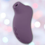 The LELO Sona 3 Cruise - Plum, a curved, purple silicone device with a circular opening at one end, is shown against a softly blurred background with pink and white bokeh lights.
