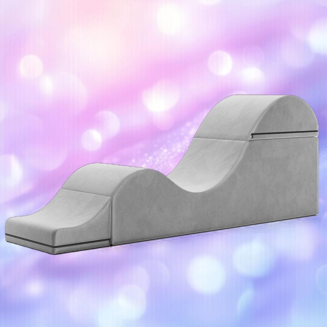 The Liberator Aria Chaise - Grey by Liberator features a curvy, wave-like design and modern upholstery, set against a pink and purple bokeh background, blending discreet sex furniture elegance with contemporary style.