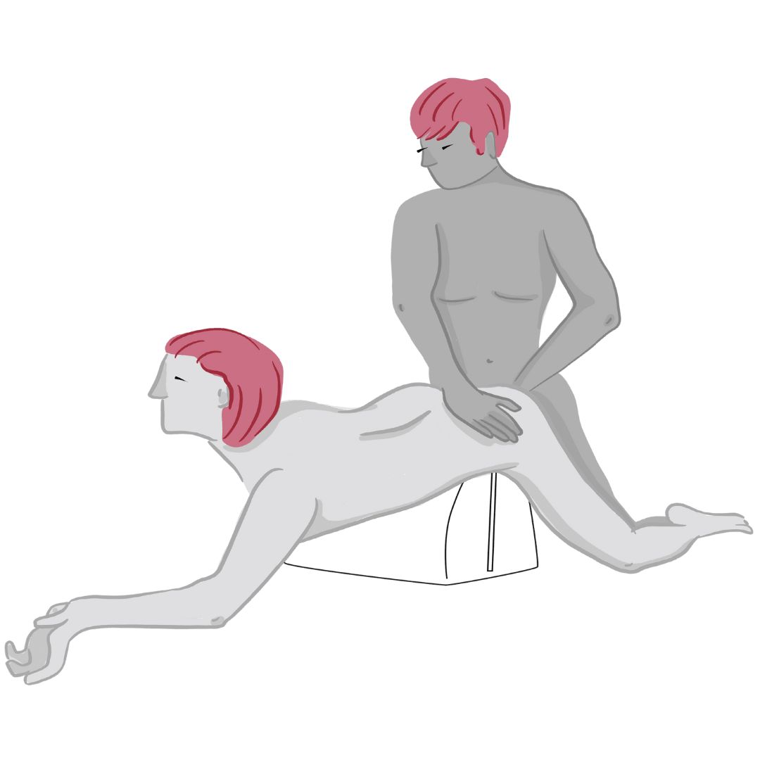 Two nude figures with pink hair are engaged in a sexual position. One lies face down on a teal Liberator BonBon Microvelvet cushion by Liberator, while the other kneels behind, holding their partner’s hip. The background is plain white.