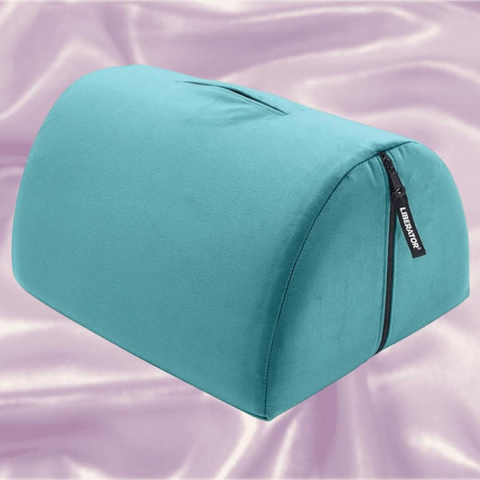 A teal Liberator Bonbon Microvelvet pillow by Liberator, dome-shaped with a zipper and handle, sits on shiny pale lavender satin—perfectly designed for hands-free solo play. 1080