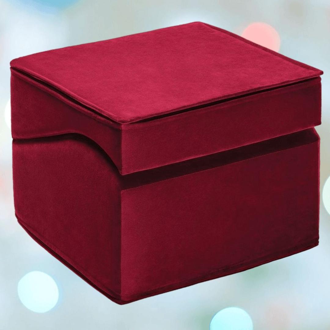 A square, deep red Liberator Flip Ramp MicroVelvet - Merlot ottoman by Liberator, with a plush texture, stands out against a blurred background with pastel-colored lights.