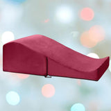 A wedge-shaped Liberator Flip Ramp MicroVelvet - Merlot cushion by Liberator rests against a pastel backdrop with soft, blurred lights, showcasing its stylish and versatile design as modern sex furniture.