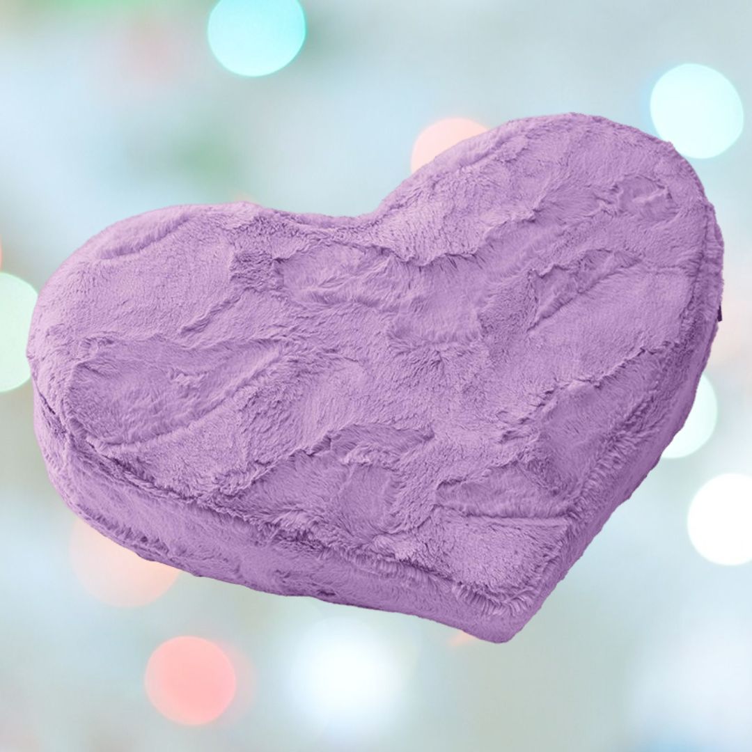 The Liberator Heart Wedge Fur in purple by Liberator features a plush, fuzzy texture and is displayed against a blurred backdrop of pastel-colored lights.